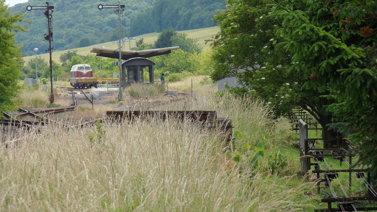 EBS 118 719-4 mit Flachwagen Richtung Naumburg, am 16.06.2015 bei der Durchfahrt in Lauch. Auf dem Bild zu erkennen sind die noch übriggebliebenen zwei durchgehenden Gleise. Von den links liegenden Weichen ist nur noch eine in Funktion, nämlich 
vom Überholgleis zum durchgehenden Hauptgleis. Rechts unten die altbewährte Stellwerkstechnik vom Stellwerk Lw, zur Betätigung der beiden Ausfahrsignale und der einen Weiche. Hinter dem Gebüsch lugt die Hütte hervor in der sich die elektronische 
Stellwerkstechnik befindet, welche das ehemals zweite Lauchaer Stellwerk Lo ersetzt 
und jetzt von Lw aus mit bedient wird. Laucha hatte mal 26 Weichen, bis in die 1960er Jahre noch vorhanden. Ganz links ist noch die der Rest von Stellwerk Lo geringfügig zu erkennen. (Foto: Günther Göbel)
